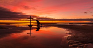 man kneeling before cross on beautiful beach sunset