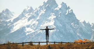 Person standing on a fence in front of a mountain praising the Lord