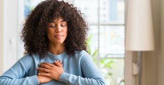 woman with closed eyes and prayer hands over chest sitting by windown