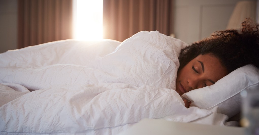 woman laying in bed eyes closed sleeping as sun peeks through window