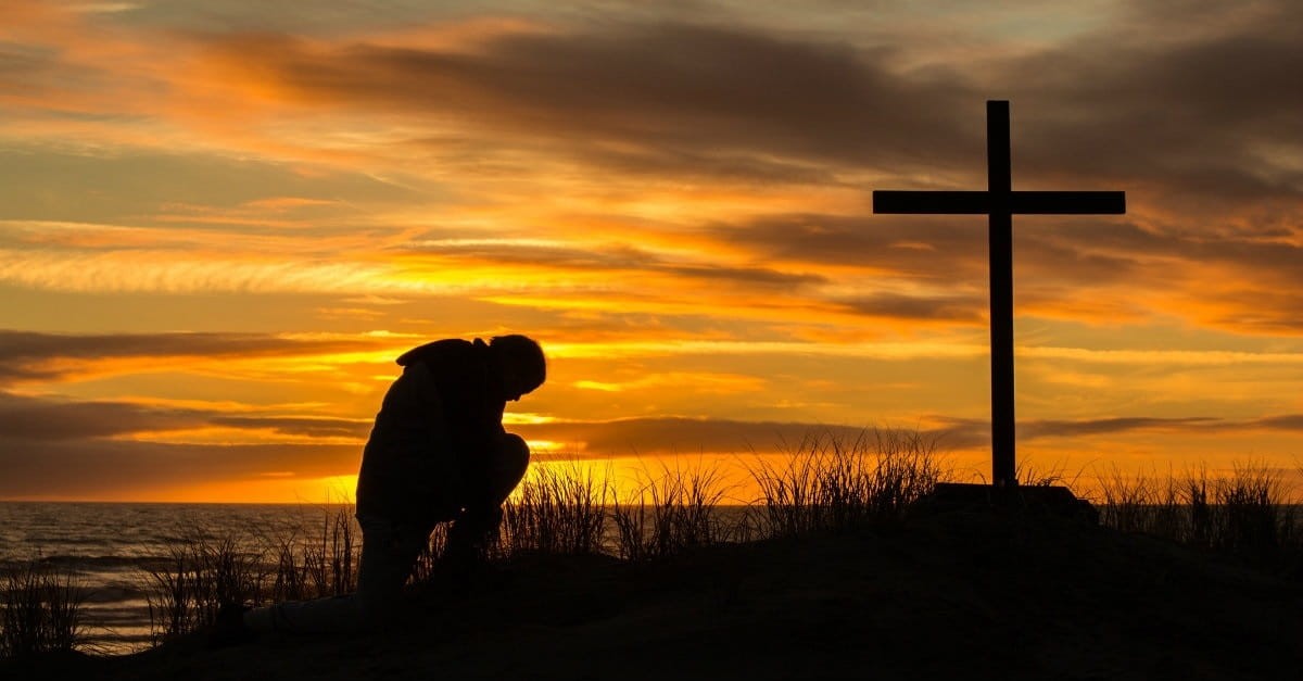 silhouette of man kneeling before a cross in a sunset lit field