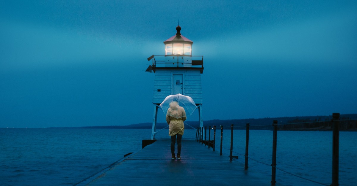 person in raincoat under umbrella walking down bridge toward lighthouse at night