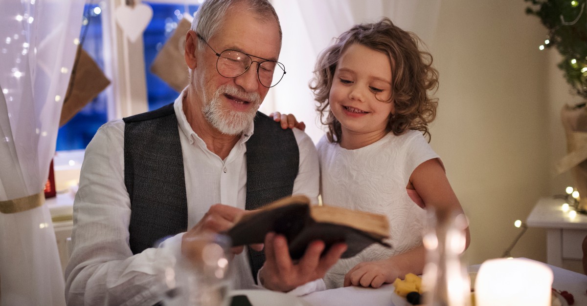 senior man reading Bible with granddaughter with little white lights in background