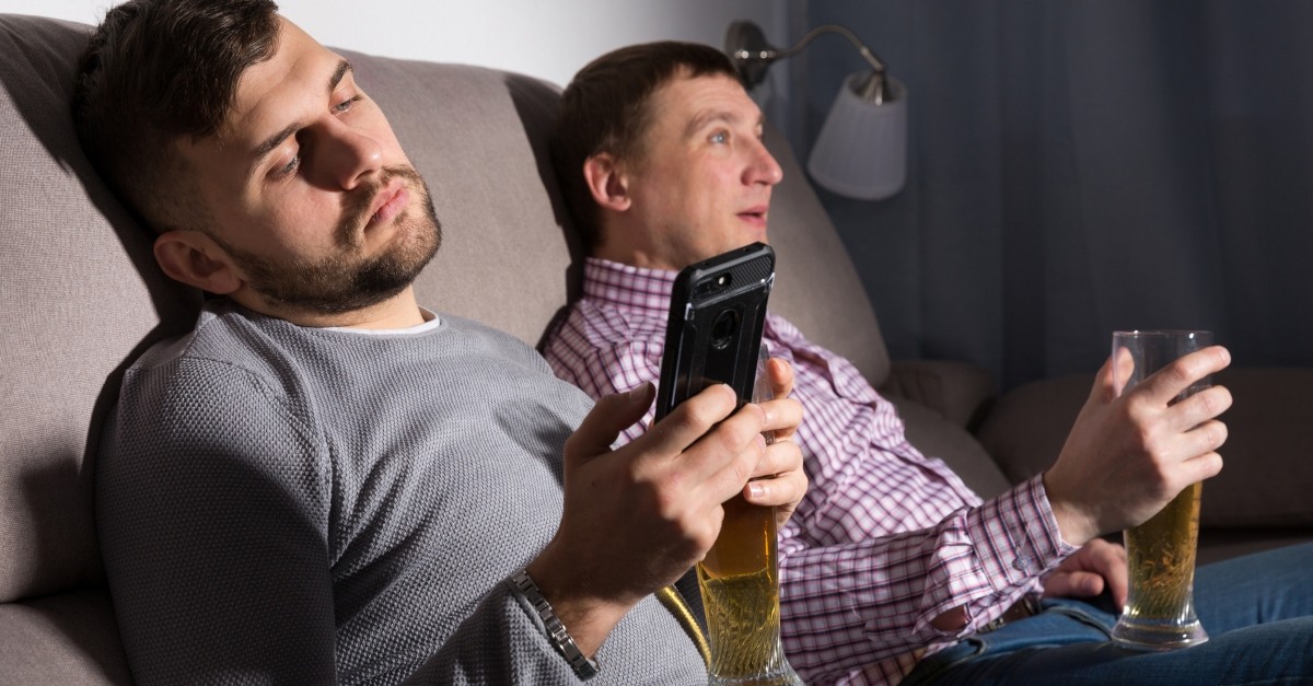 two men lazing on couch with beers one watching TV the other staring at smartphone