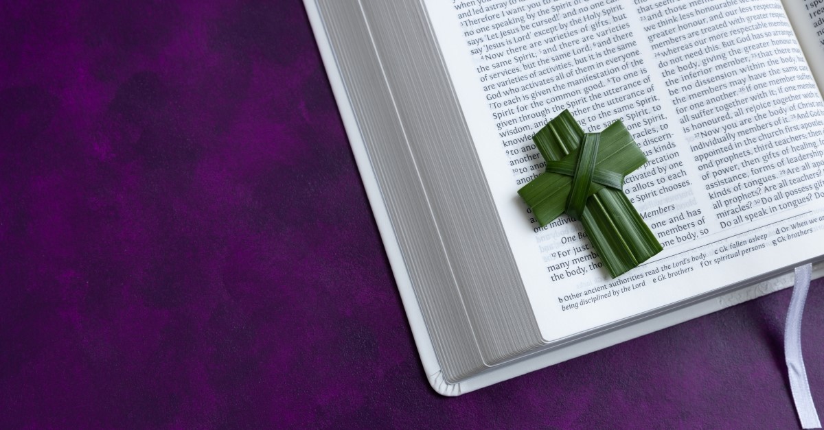 Small cross made from a palm leaf laying on an open white bible on a dark purple background shot from above, bible verses lent