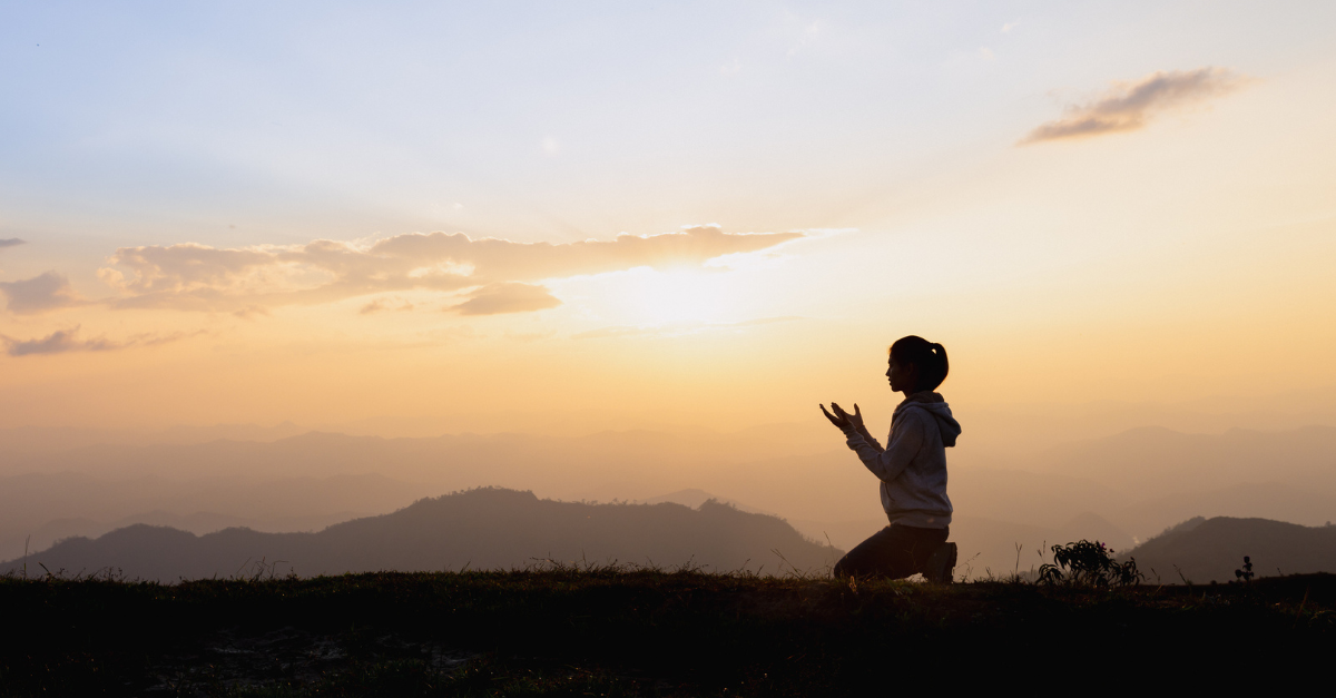 Silhouette of a woman, knelt down in a field praying at dusk.