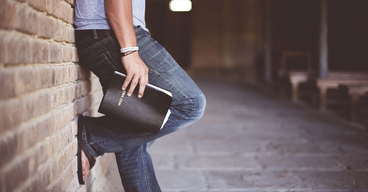 Man, leaning up against a wall outside, holding a closed Bible in his hand.