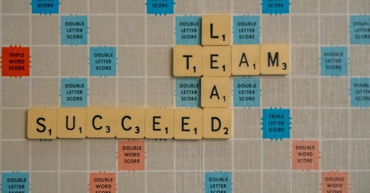 Scrabble tiles spelling “TEAM” intersecting with “LEAD” and “SUCCEED” on a game board, symbolizing leadership, teamwork, and success