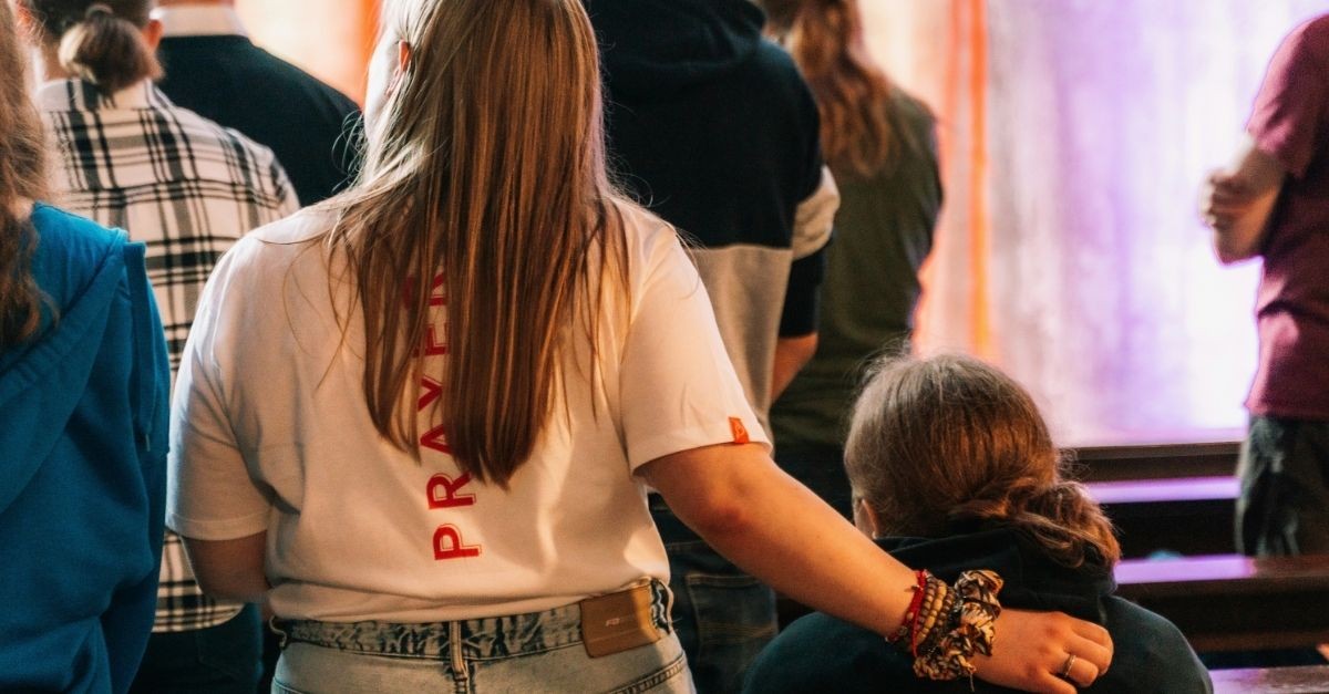 Church attendees stand together during a worship service, with one person placing an arm around another in a gesture of support, friendship, and Christian community.