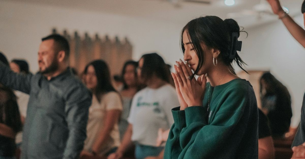 A young woman stands in prayer during a church worship service, eyes closed and hands clasped, while other congregants worship in the background.