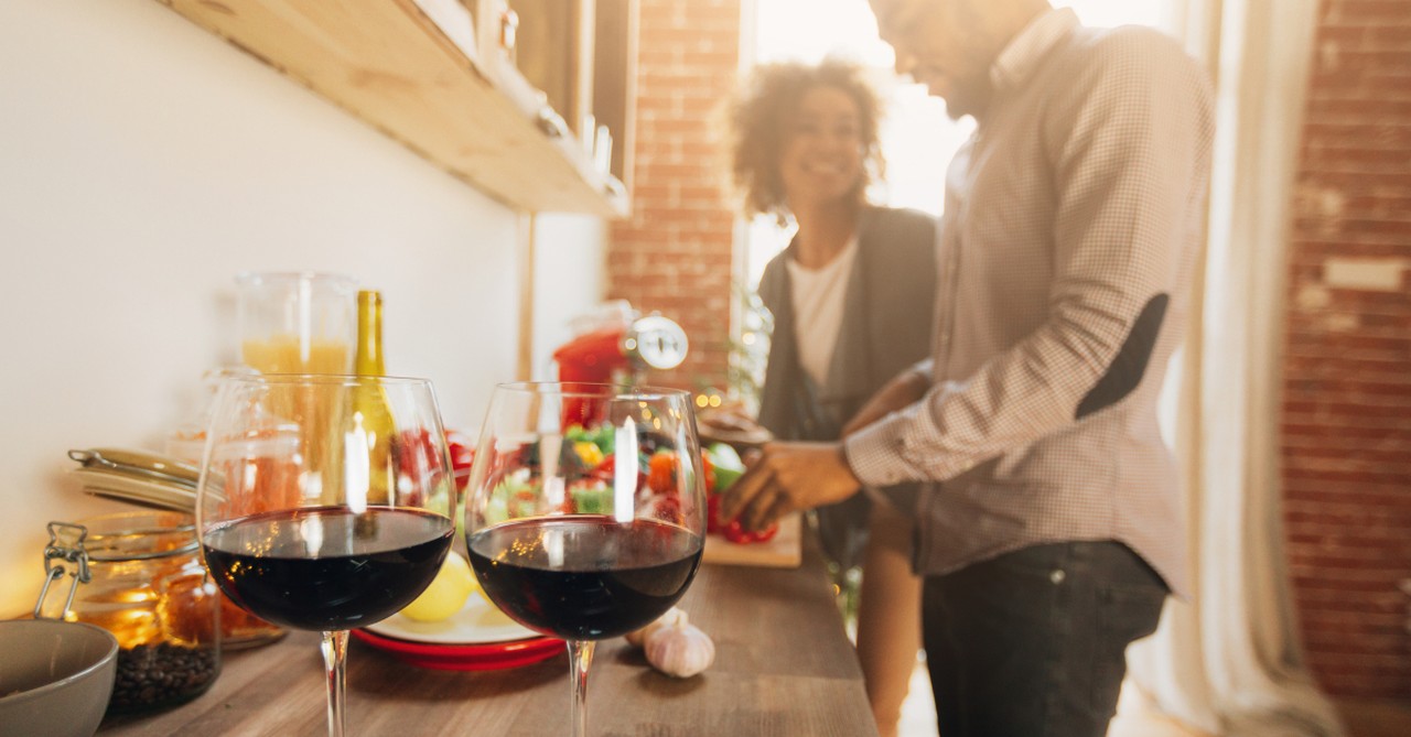 couple prepping dinner together two glasses red wine in foreground