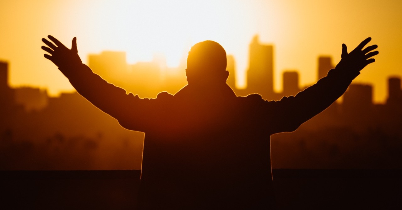 man with hands raised at sunrise over city