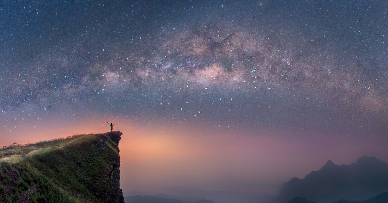 man standing on green meadow cliff looking out at stars