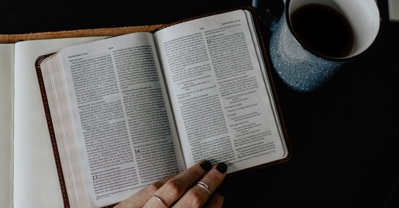 open Bible on table with hand resting on top and coffee above