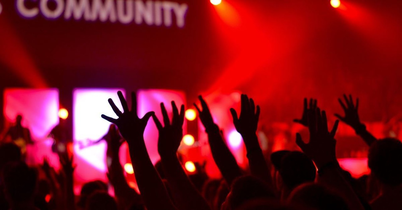 hands raised in audience in worship against red lights background with community printed on sign