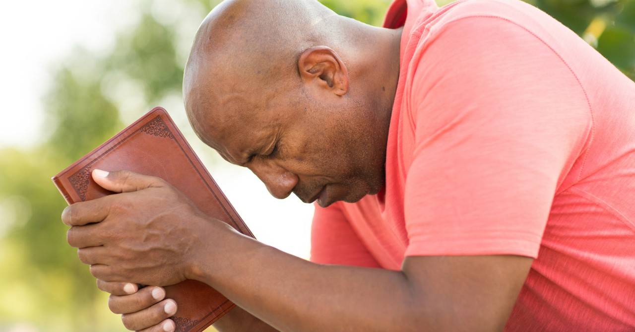 side view of man holding bible with head down eyes closed in prayer