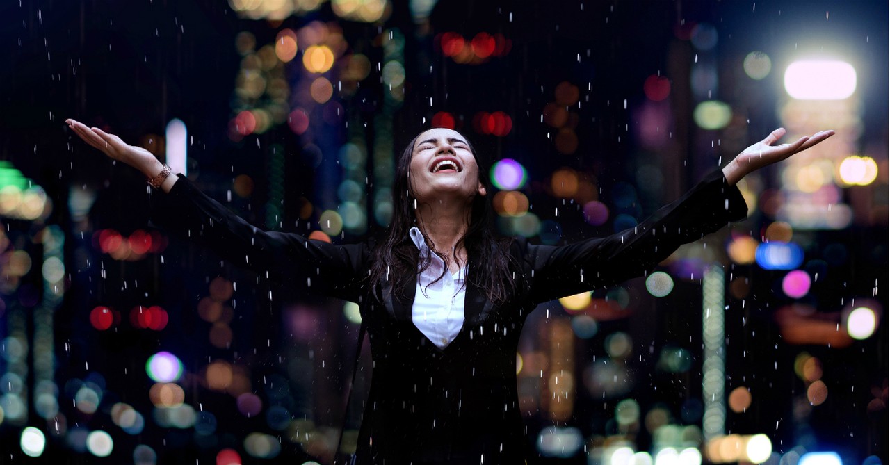 woman in black suit at night face up toward rain smiling