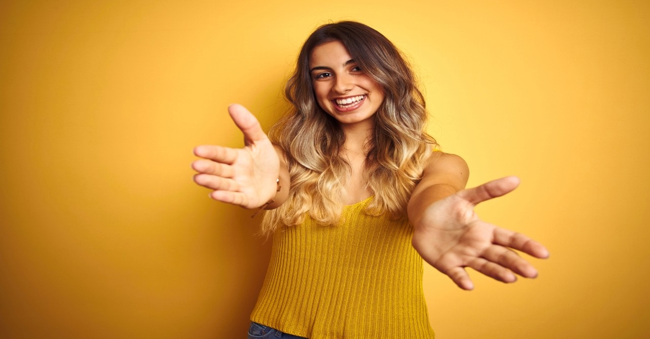 Young woman over yellow background looking at the camera smiling with open arms for hug.