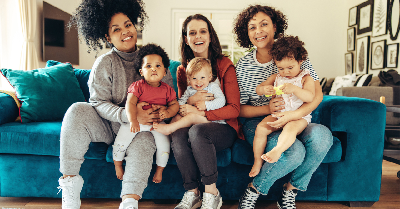 Three mom friends and their little kids sitting on a couch laughing together.