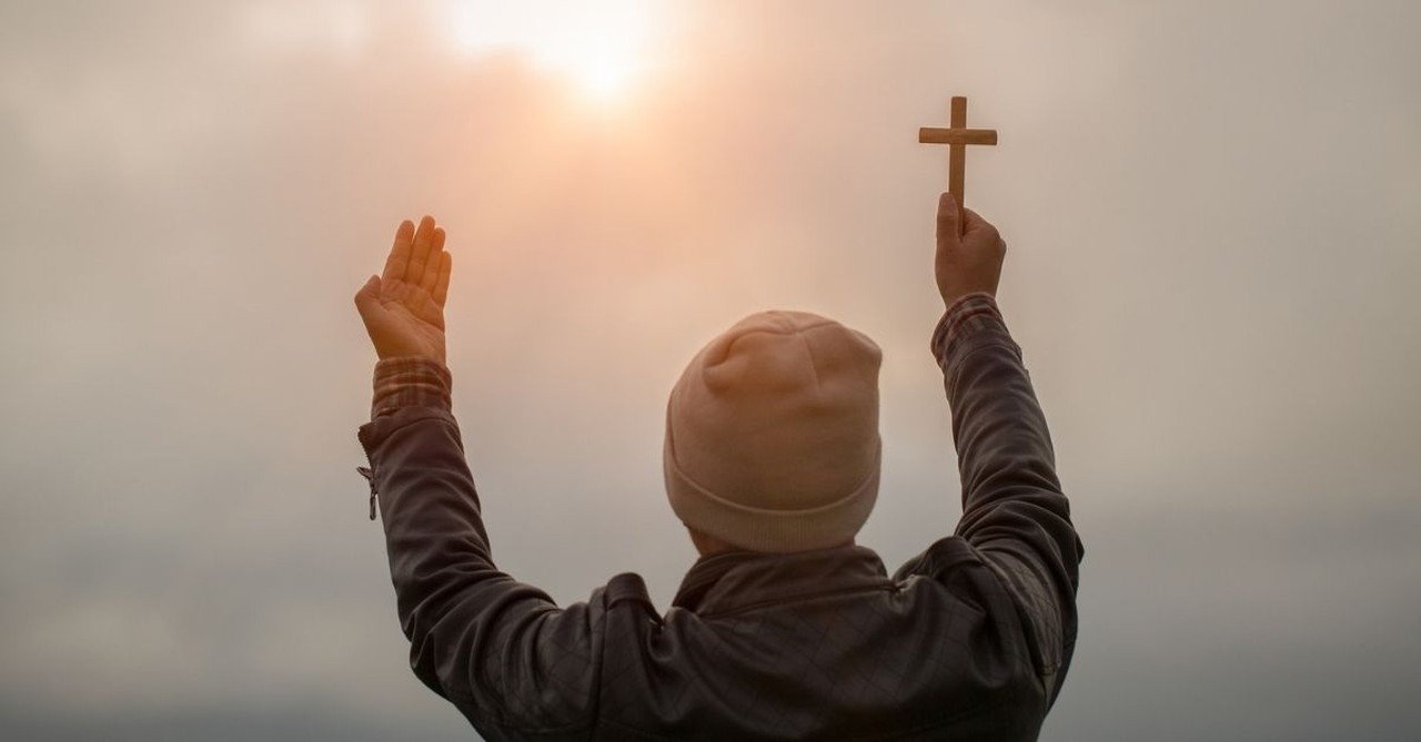 Man praying with cross in hand, looking towards an opening of light in the sky;