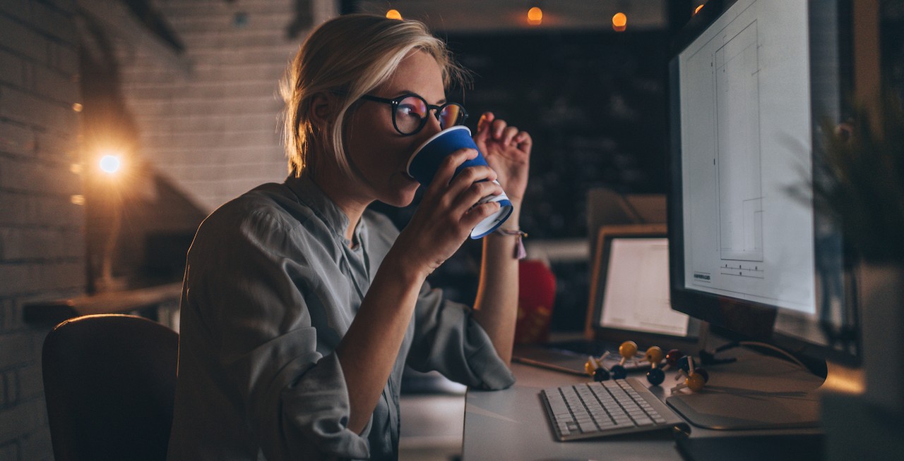 Woman working late in office at job at night drinking coffee