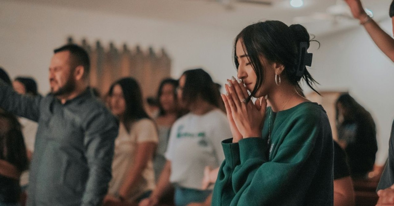 A young woman stands in prayer during a church worship service, eyes closed and hands clasped, while other congregants worship in the background.