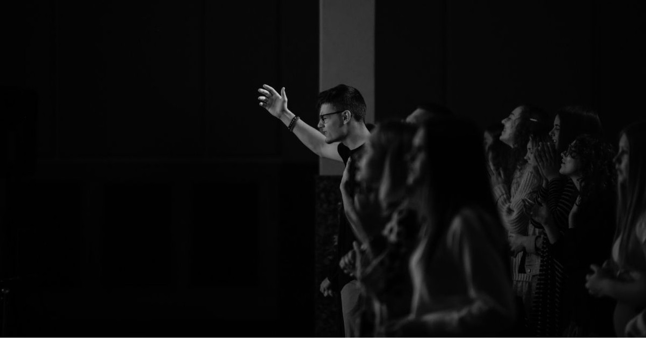 A group of young adults worships together in a darkened church sanctuary, with one person raising a hand in praise during a worship service.