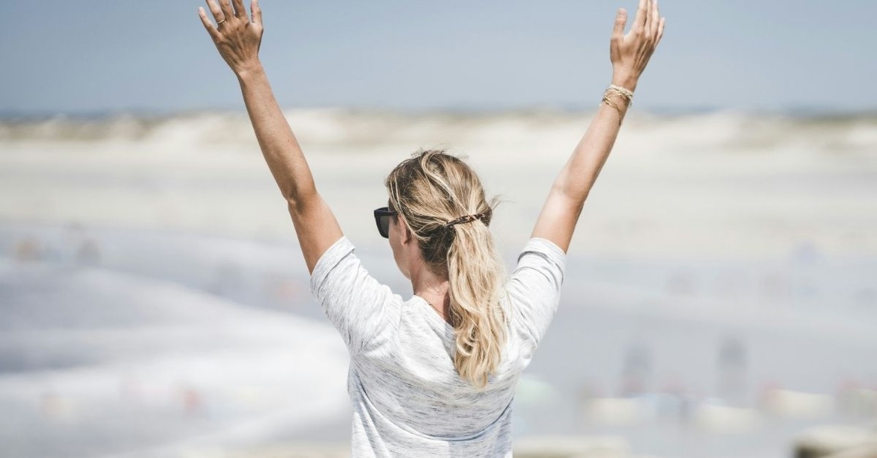 A woman stands outdoors with her arms raised toward the sky, expressing gratitude, praise, and joyful worship in an open natural setting.