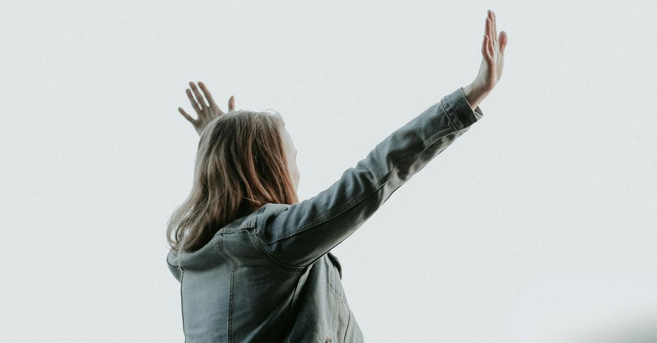 A woman stands outdoors with both hands raised toward the sky in worship and prayer, expressing surrender, praise, trust, and wholehearted devotion to God.