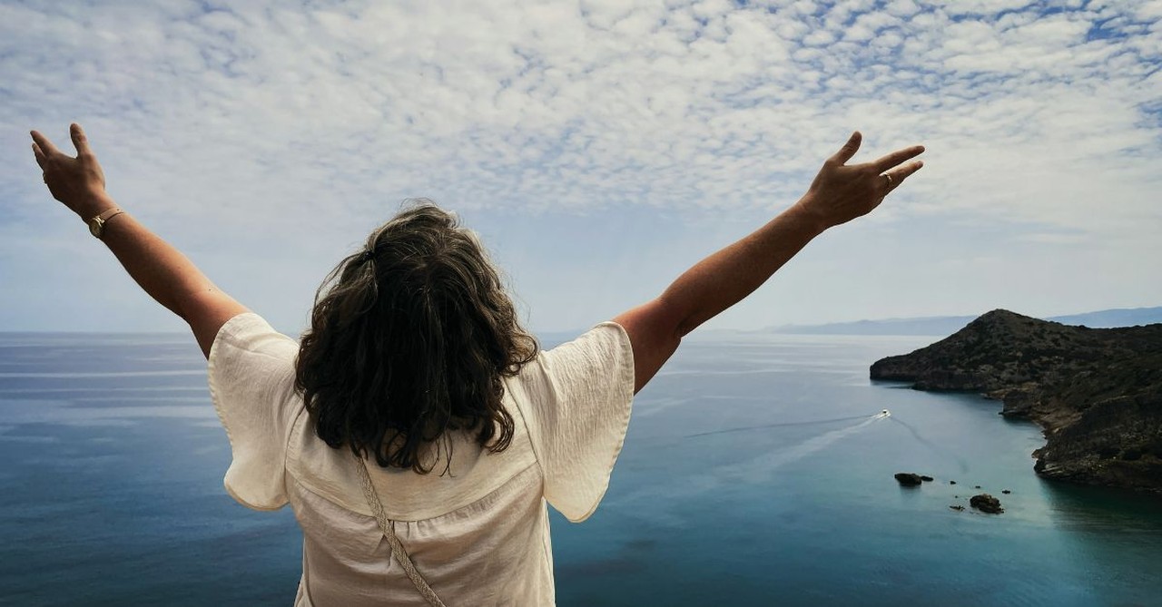 A woman stands on a scenic coastal overlook with her arms raised toward the sky, gazing over the ocean in an expression of praise, gratitude, freedom, and worship as she takes in God's creation.