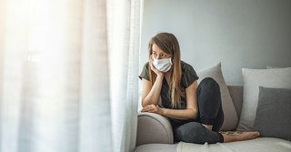 woman wearing mask sitting on sofa looking out window