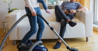 Wife vacuuming while her husband sits on the couch.