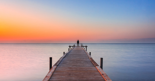 Person watching the sunrise at the end of a long dock over the water.
