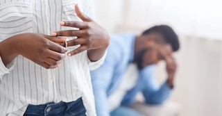 A woman removing her wedding ring while her husband looks despondent in the background