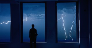 Man standing at a window, watching a lightning storm