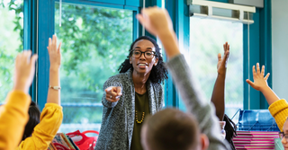Teacher in her classroom, children with hands raised; a message for teachers this summer.