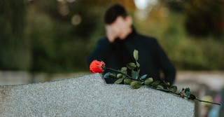 A man grieving at a tombstone with a single red rose on top of it