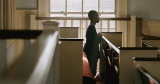 A person stands alone in a church pew, praying quietly as sunlight streams through large windows, creating a reflective and contemplative atmosphere.