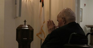 An elderly man sits in a church sanctuary with his hands raised in prayer, facing a cross and baptismal font in a quiet moment of worship and reflection.