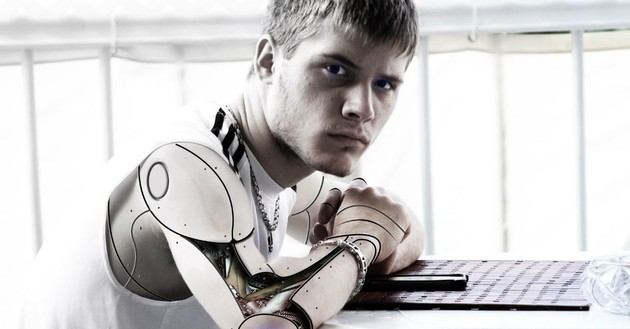 man turning head to face camera, leaning on desk with artificial arm