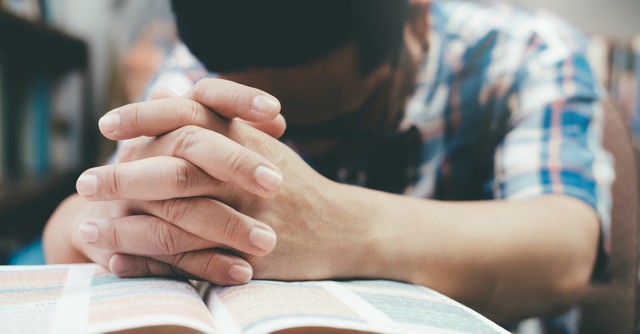 man with head down hands folded on open Bible praying
