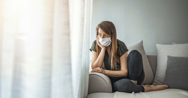 woman wearing mask sitting on sofa looking out window