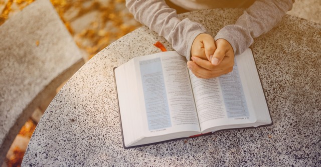 prayer hands resting on open Bible at table outside