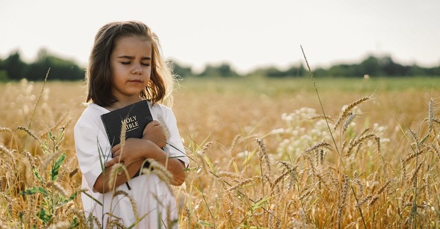 little girl holding Bible outside in a field with her eyes closed
