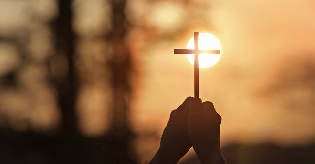 A person holding up a small cross to the setting sun