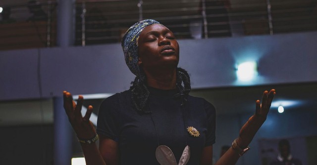 A woman stands in worship with her eyes closed and hands lifted in prayer inside a church, expressing surrender, reverence, trust, and wholehearted devotion to God.