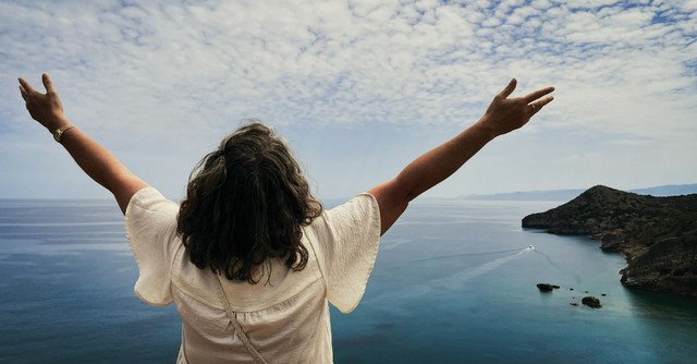 A woman stands on a scenic coastal overlook with her arms raised toward the sky, gazing over the ocean in an expression of praise, gratitude, freedom, and worship as she takes in God's creation.