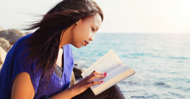 woman reading bible in purple shirt by ocean