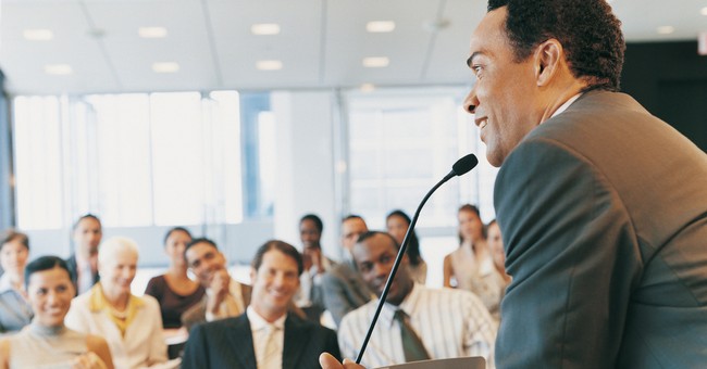 man speaking at podium with microphone to audience smiling