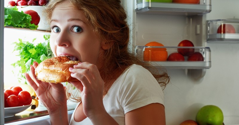 woman looking guilty eating snack from refrigerator late at night